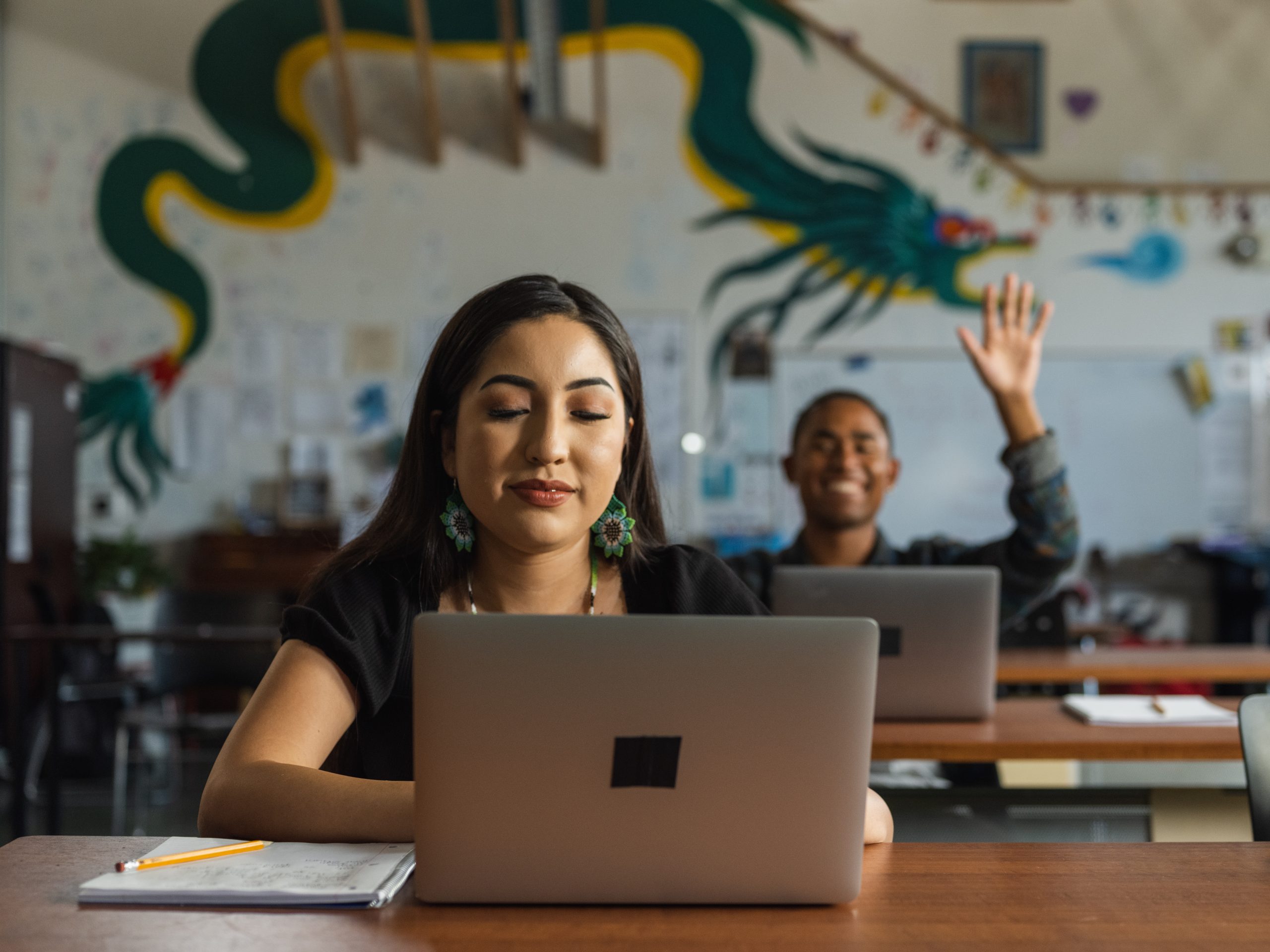 A woman is sitting at a desk, looking at her laptop, while a man in the background raises his hand.